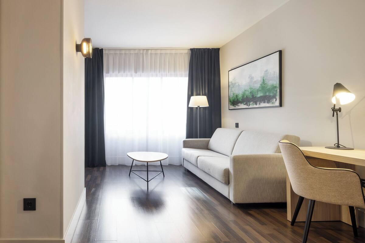 A hotel room with a bed, a desk, a chair, a small table, and a painting on the wall.