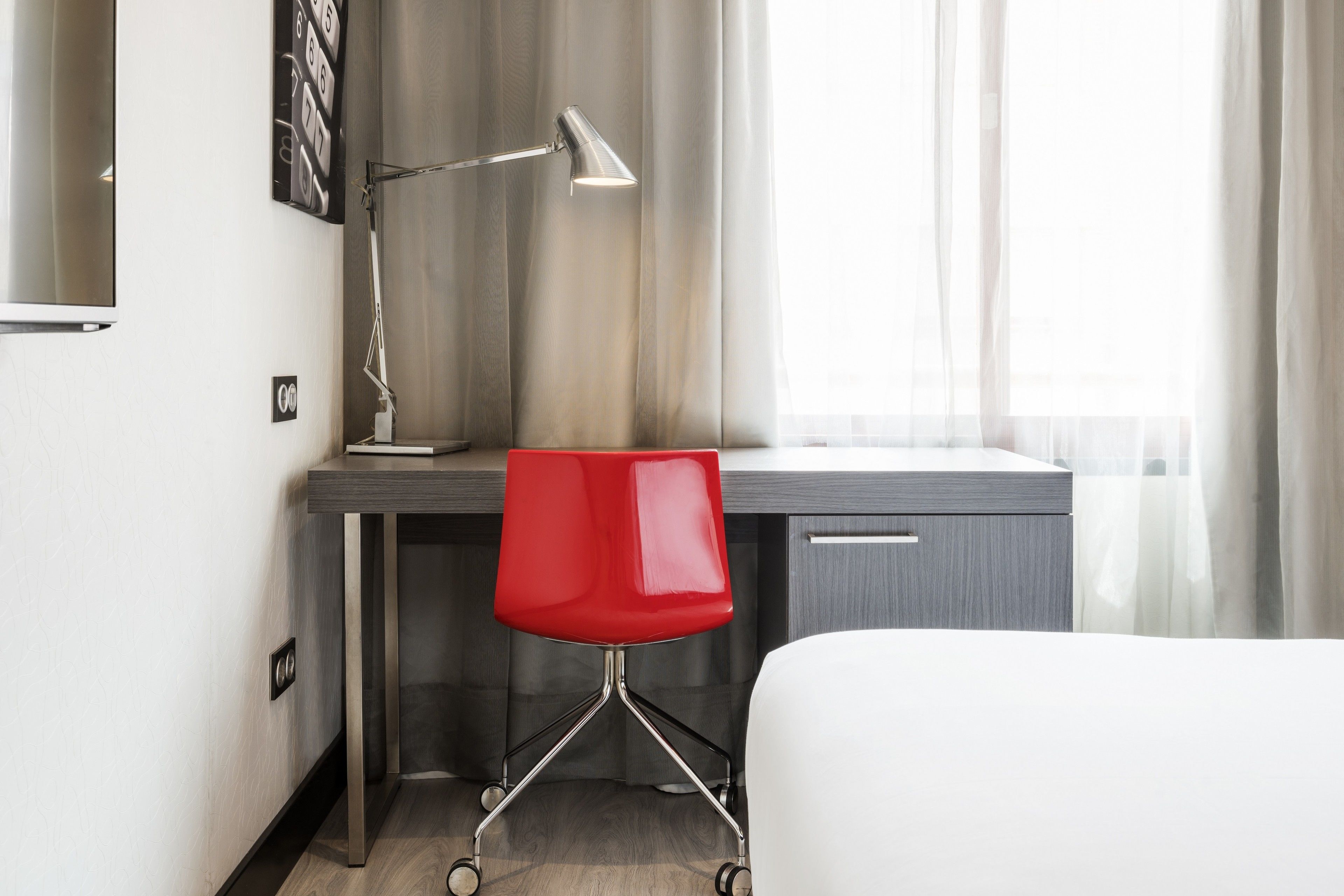 A hotel room with a desk, a red chair, a lamp, and a bed.