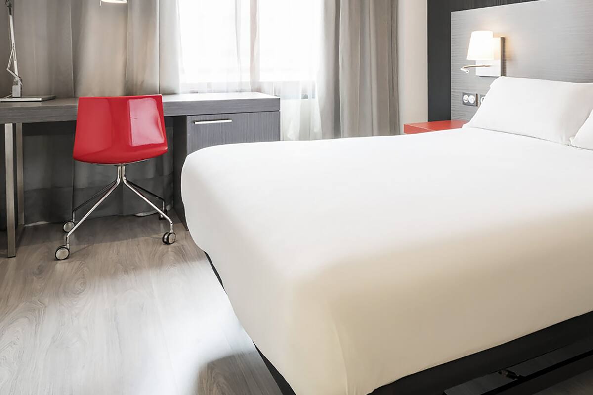 A hotel room with a large bed, a desk with a chair, and a nightstand with a lamp.