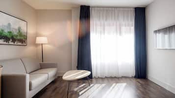 A modern hotel room with a sofa, a small round table, a floor lamp, and a large window with curtains.