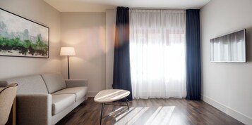 Ein modernes Hotelzimmer mit einer Couch, einem kleinen runden Tisch, einer Stehlampe und einem großen Fenster mit Vorhängen.