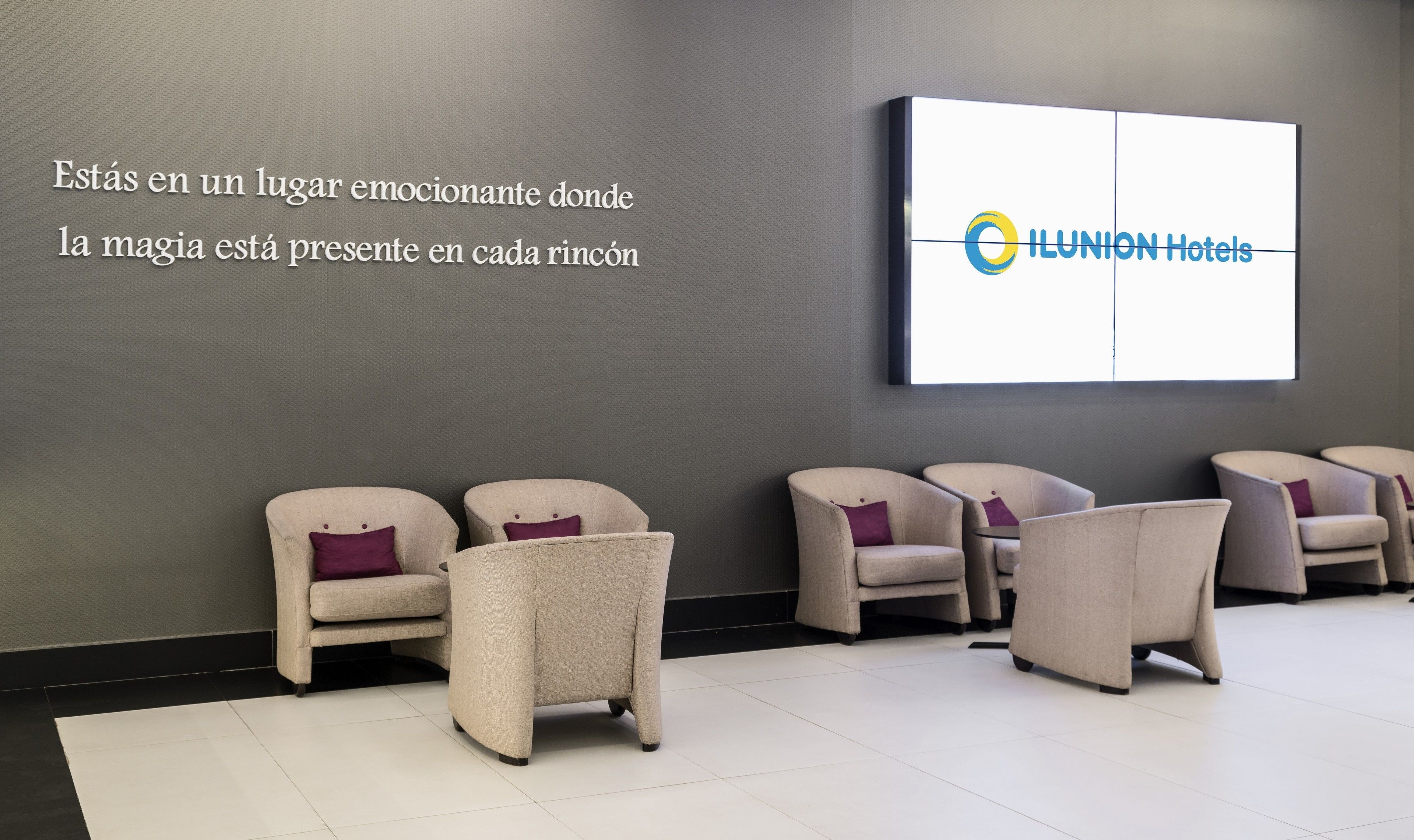Un hall d’hôtel au design moderne, comprenant un espace salon avec des fauteuils et un grand écran fixé au mur, sur lequel est affiché le logo de l’hôtel.