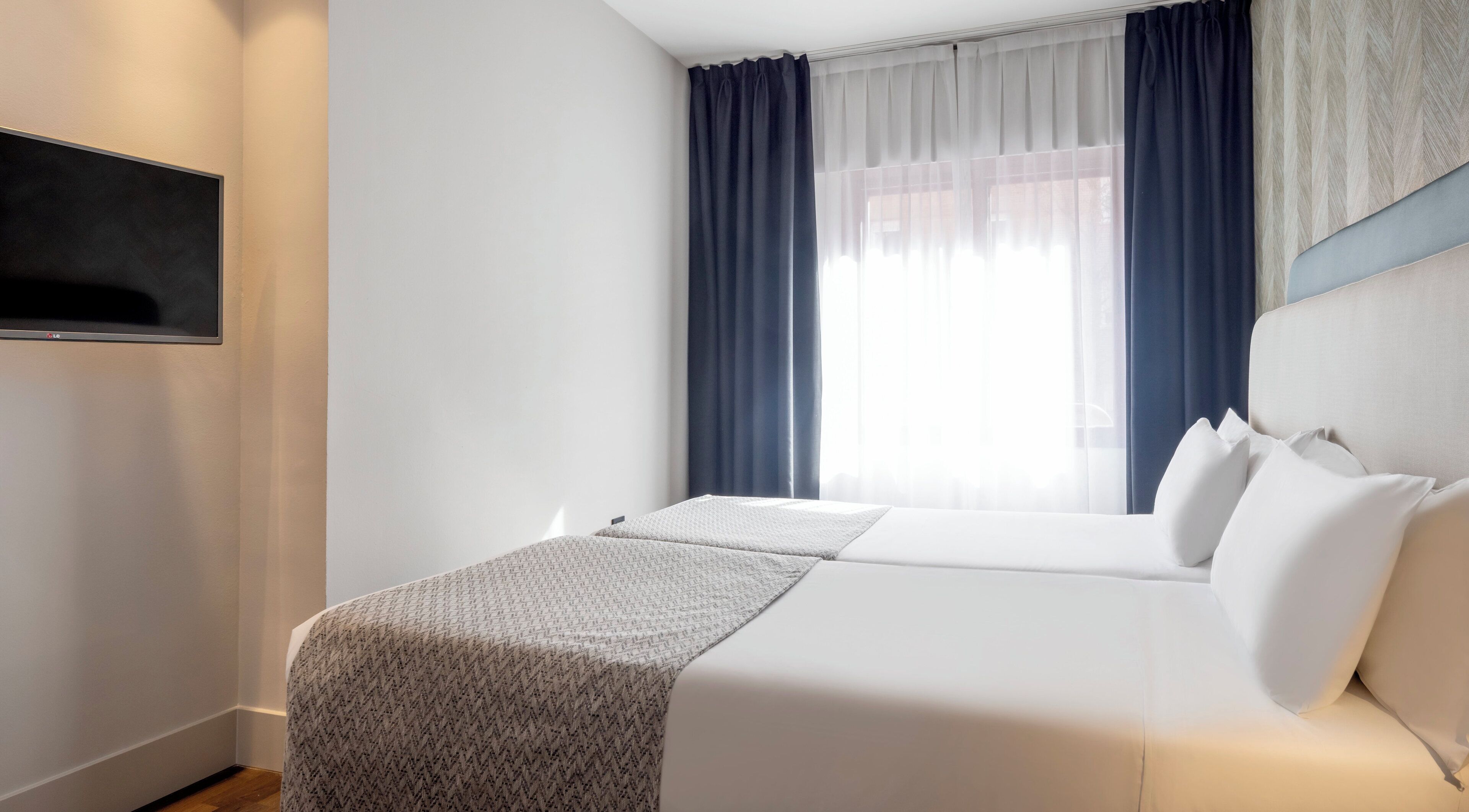 Una habitación de hotel con una cama, un televisor montado en la pared y una ventana con cortinas.