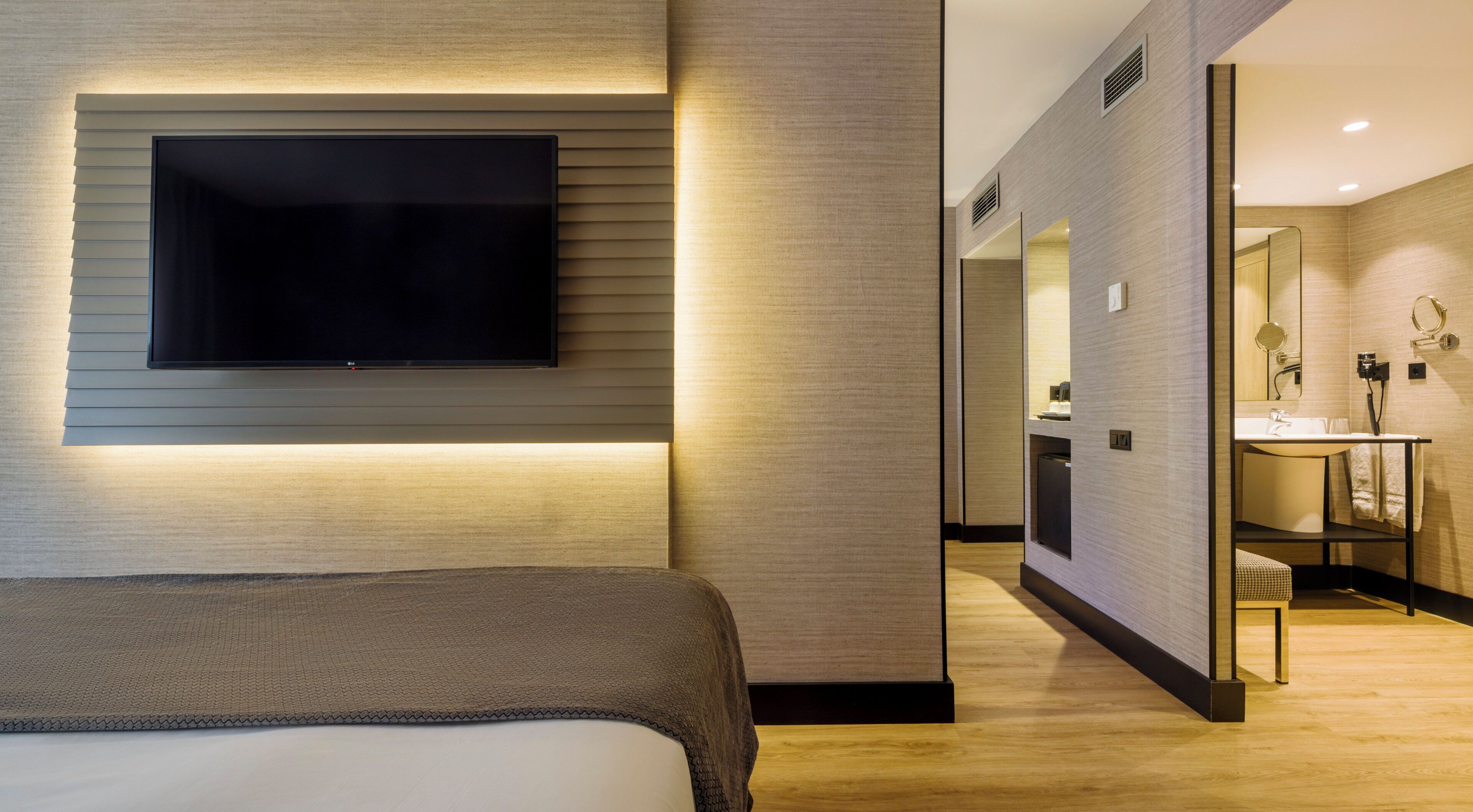 A modern hotel room with a flat-screen TV mounted on the wall, a bed with a grey headboard, and a bathroom with a mirror, sink, and towel rack.