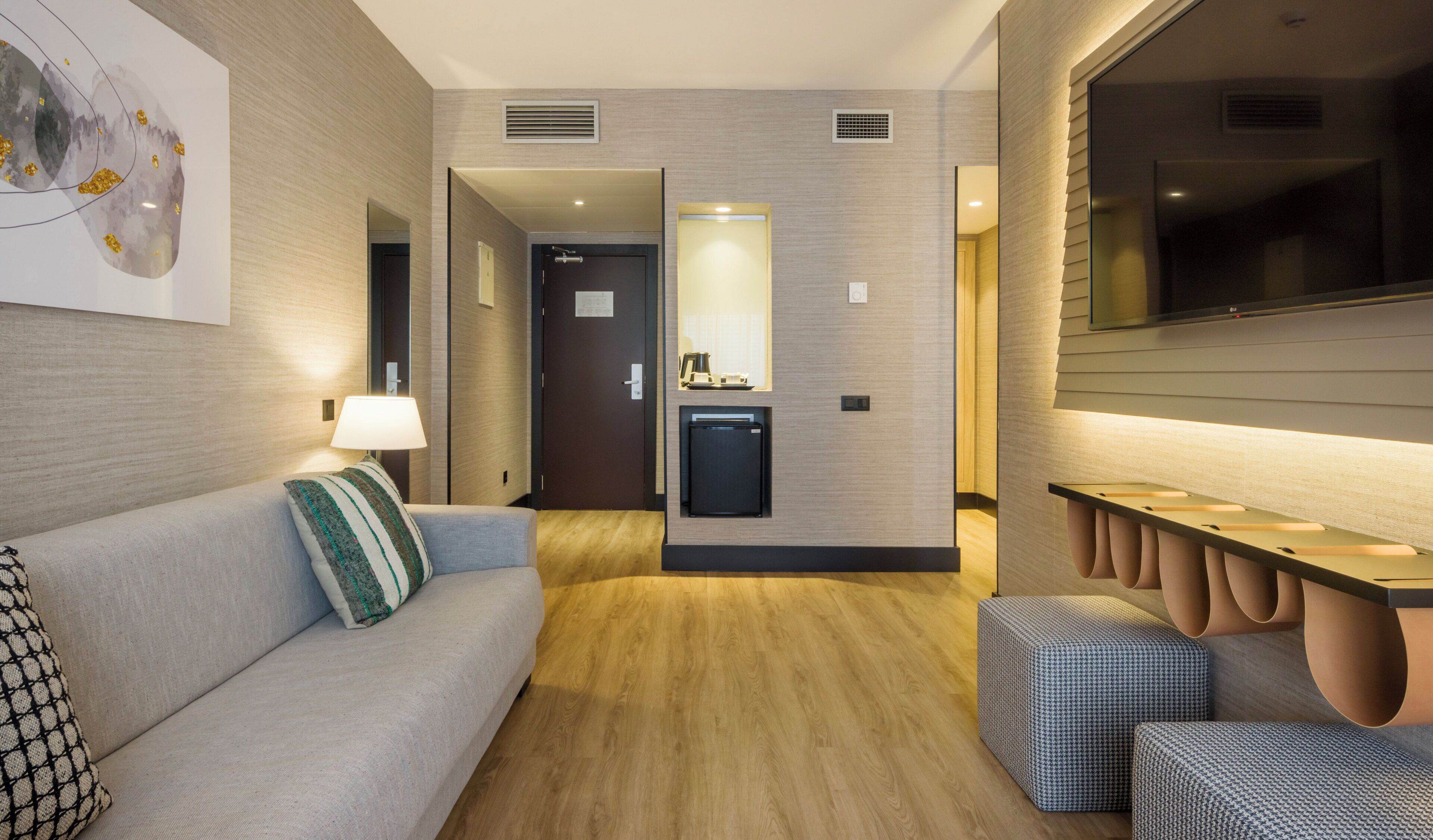 Una habitación de hotel moderna con sofá, una mesita, lámpara, mini refrigerador y televisión.