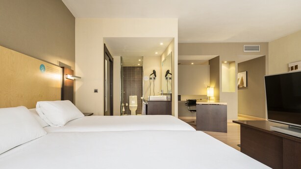 A hotel room with a large bed, a desk with a flat-screen TV, a bathroom with a shower and toilet, and a hallway leading to other rooms. - Hotel ILUNION Suites Madrid (Madrid)