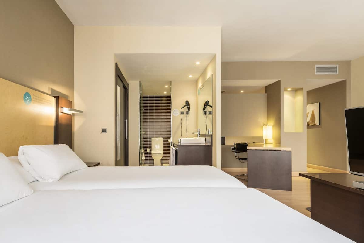 A hotel room with a large bed, a desk with a flat-screen TV, a bathroom with a shower and toilet, and a hallway leading to other rooms.
