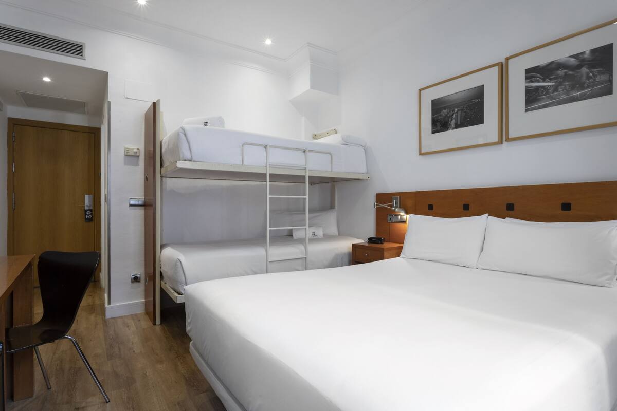 A hotel room with a bunk bed, a desk, a chair, a nightstand, and two framed pictures on the wall.