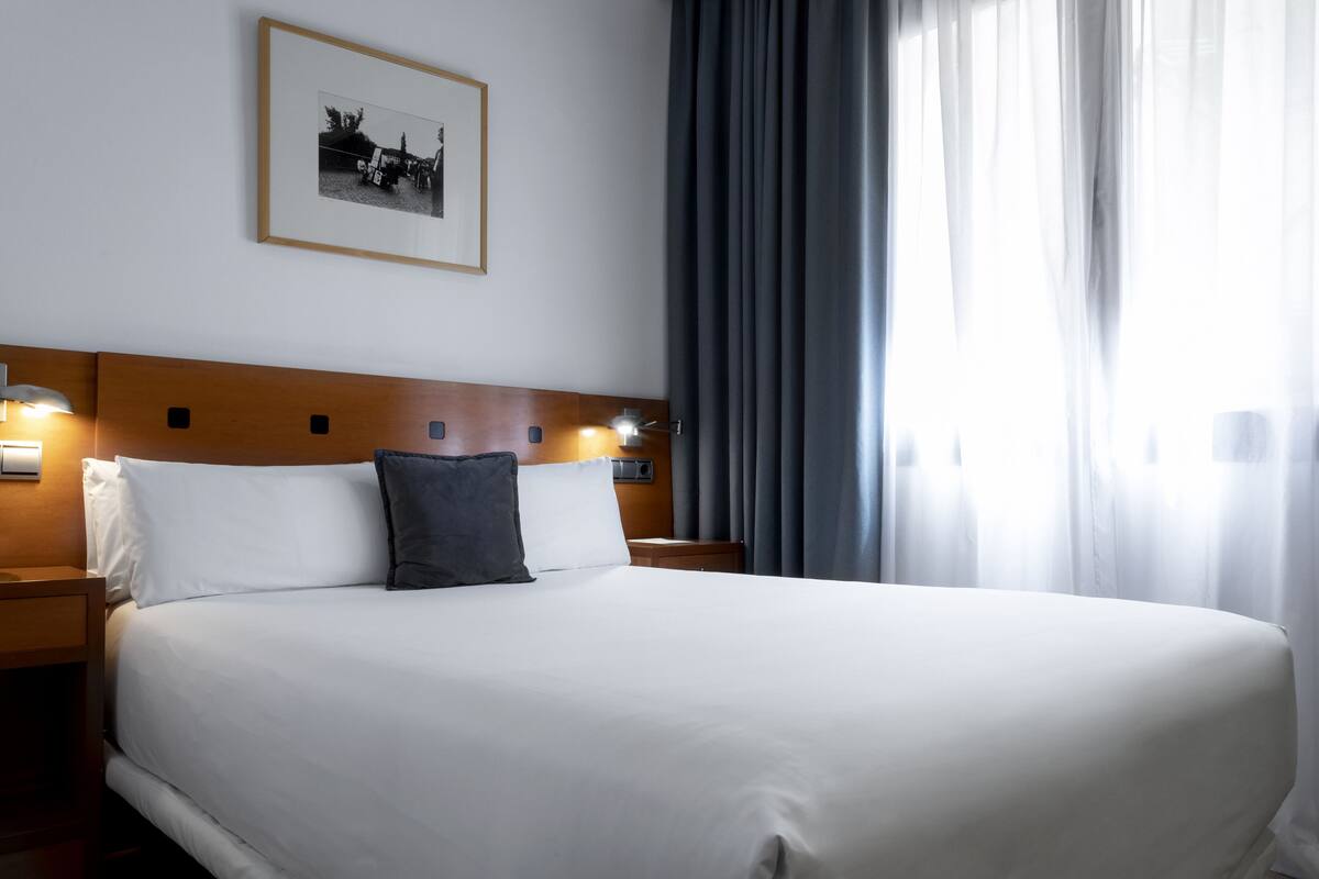 A neatly made bed with white linens and a single dark pillow, a wooden headboard, a framed picture on the wall, and a nightstand with a lamp.