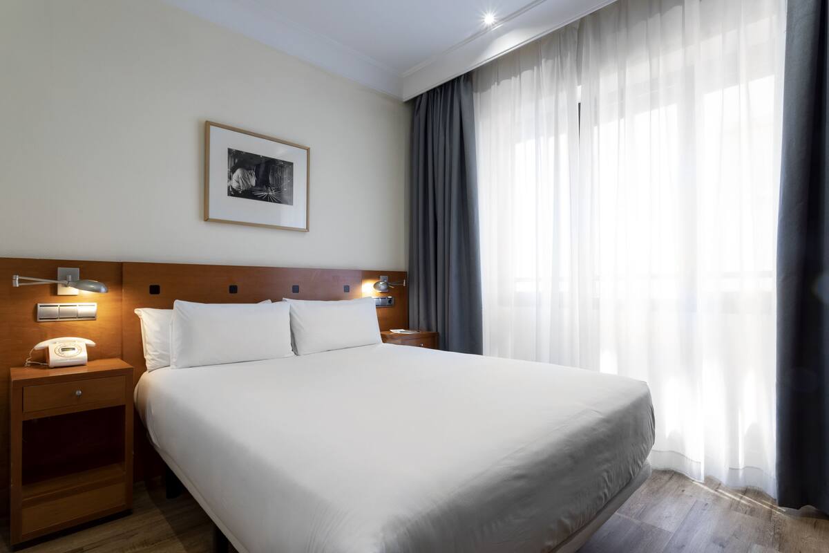 A hotel room with a large bed, a bedside table with a lamp, a framed picture on the wall, and a window with curtains.