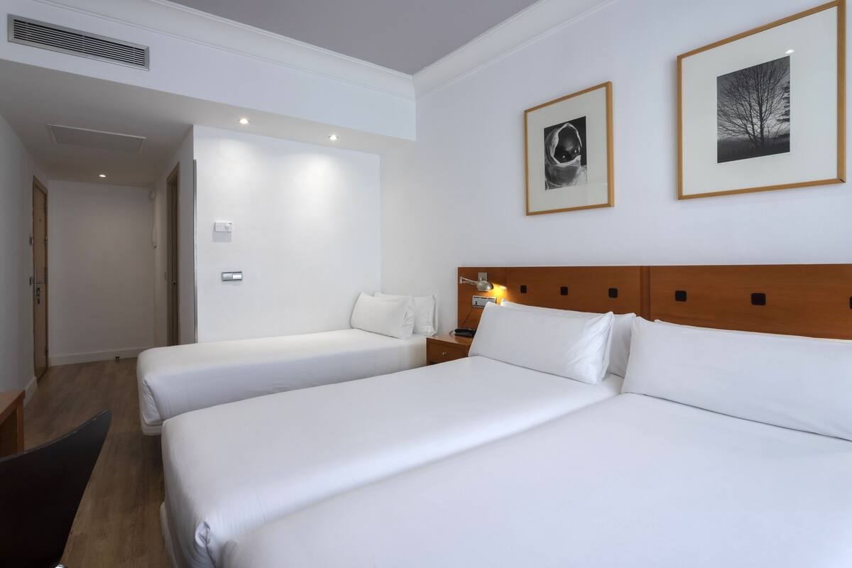 A hotel room with two single beds, a wooden headboard, a bedside table with a lamp, and framed pictures on the wall.