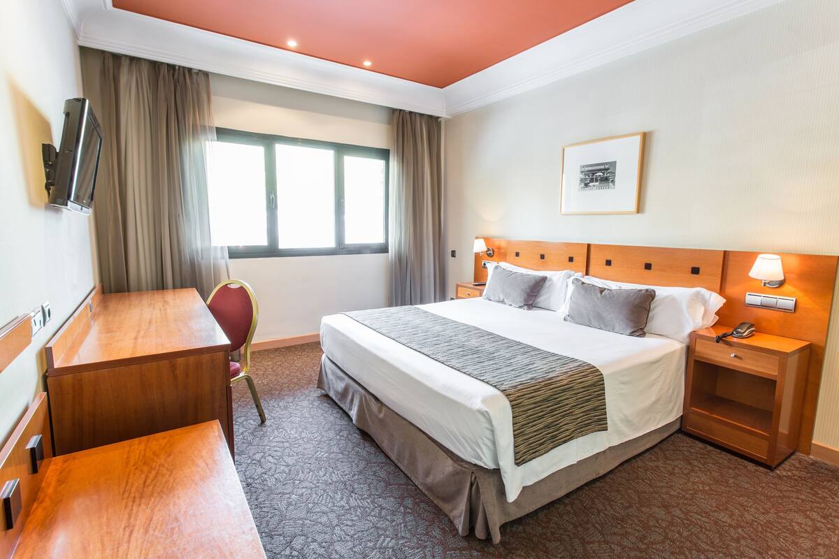 Een hotelkamer met een bed, nachtkastje, bureau, stoel en een aan de muur bevestigde televisie.