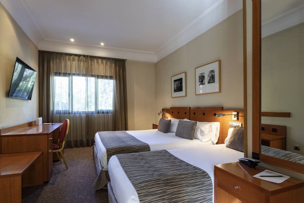Een hotelkamer met twee bedden, een bureau, een stoel, een grote spiegel en een raam met gordijnen.