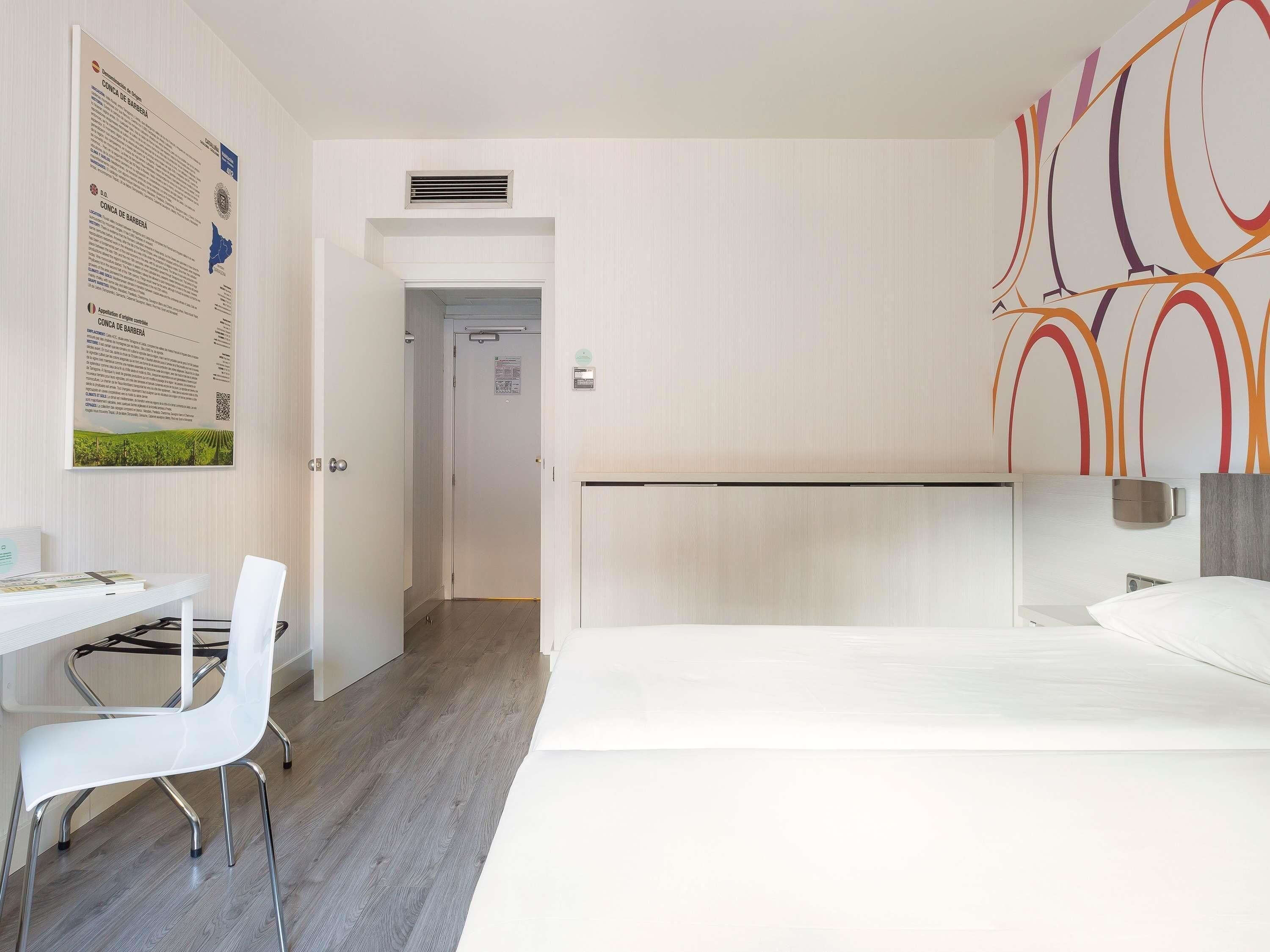 A hotel room with a bed, a desk, a chair, and a wall with a colorful abstract design.