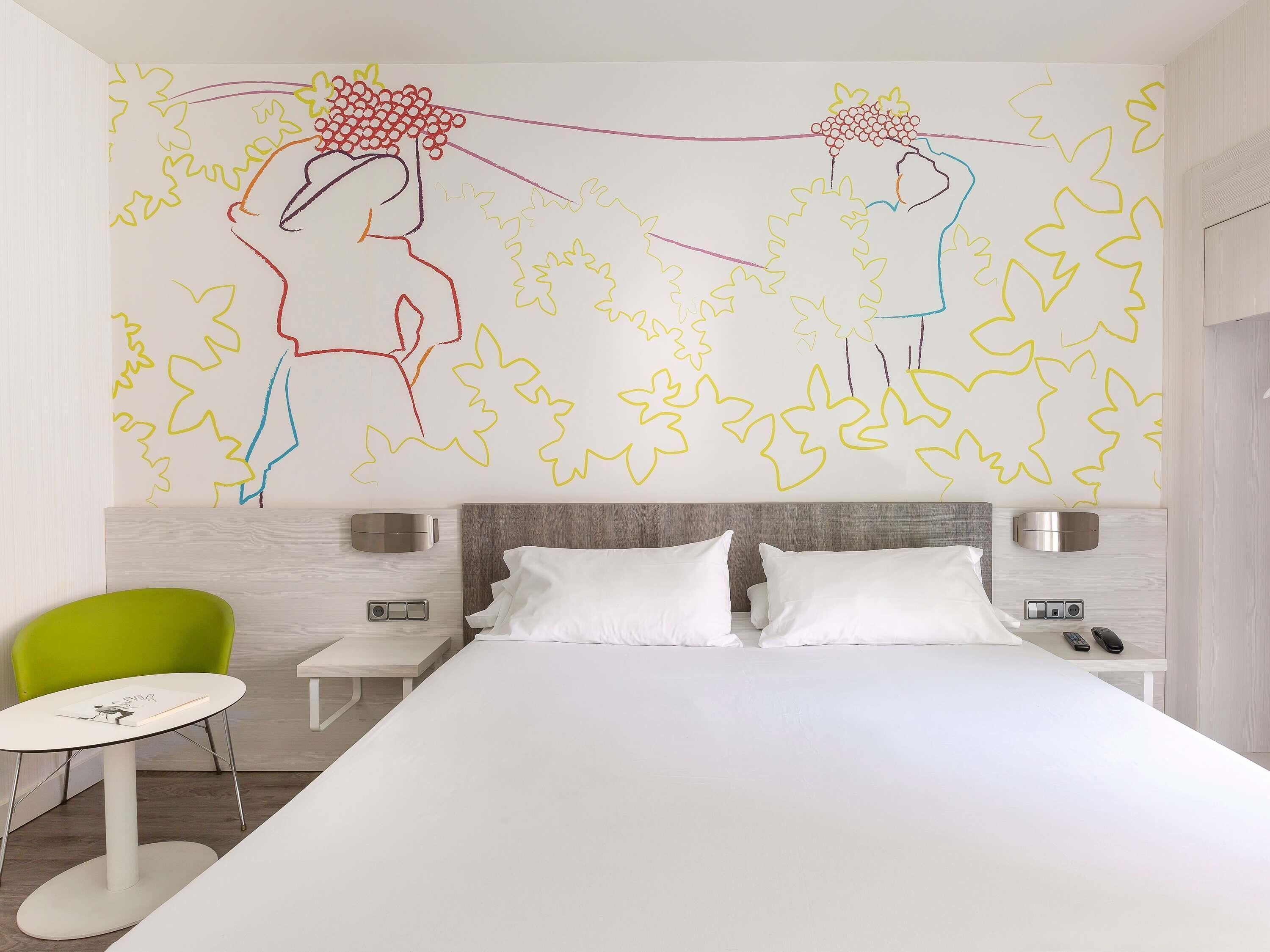 A modern hotel room with a large bed, a green chair, a small table, and a wall with abstract art.