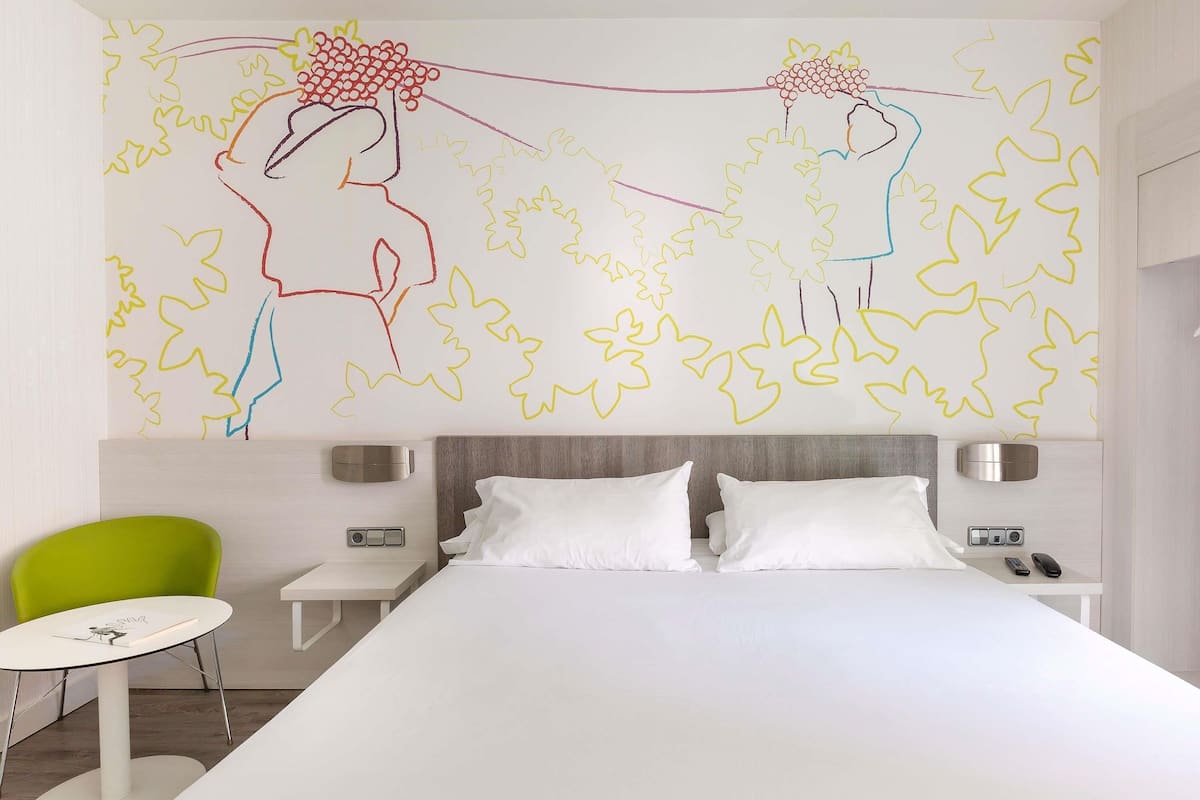 A modern hotel room with a large bed, a green chair, a small table, and a wall with abstract art.
