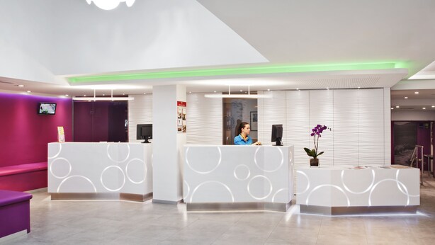 Un área de recepción moderna con una recepcionista, una zona de estar morada y una gran lámpara con forma de flor en el techo.