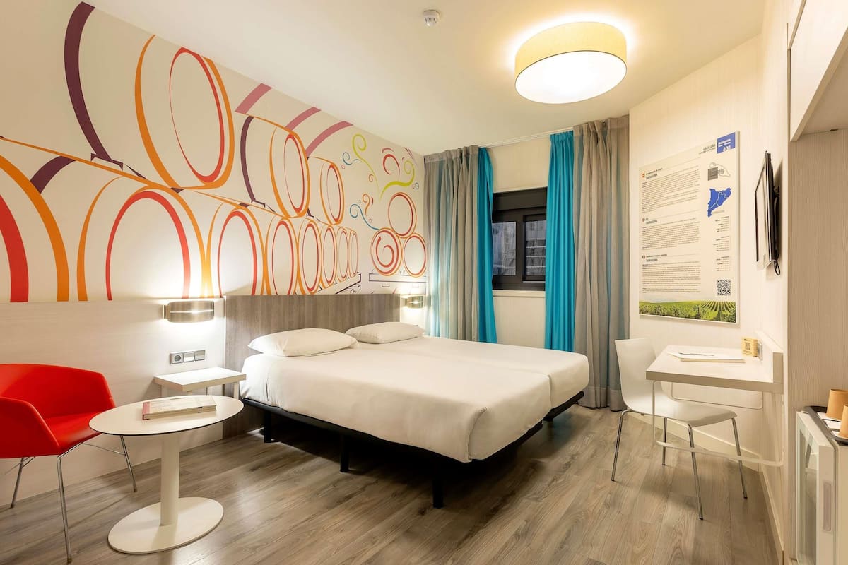 A hotel room with a bed, a desk, a chair, a red chair, a round table, and a wall with a colorful abstract design.