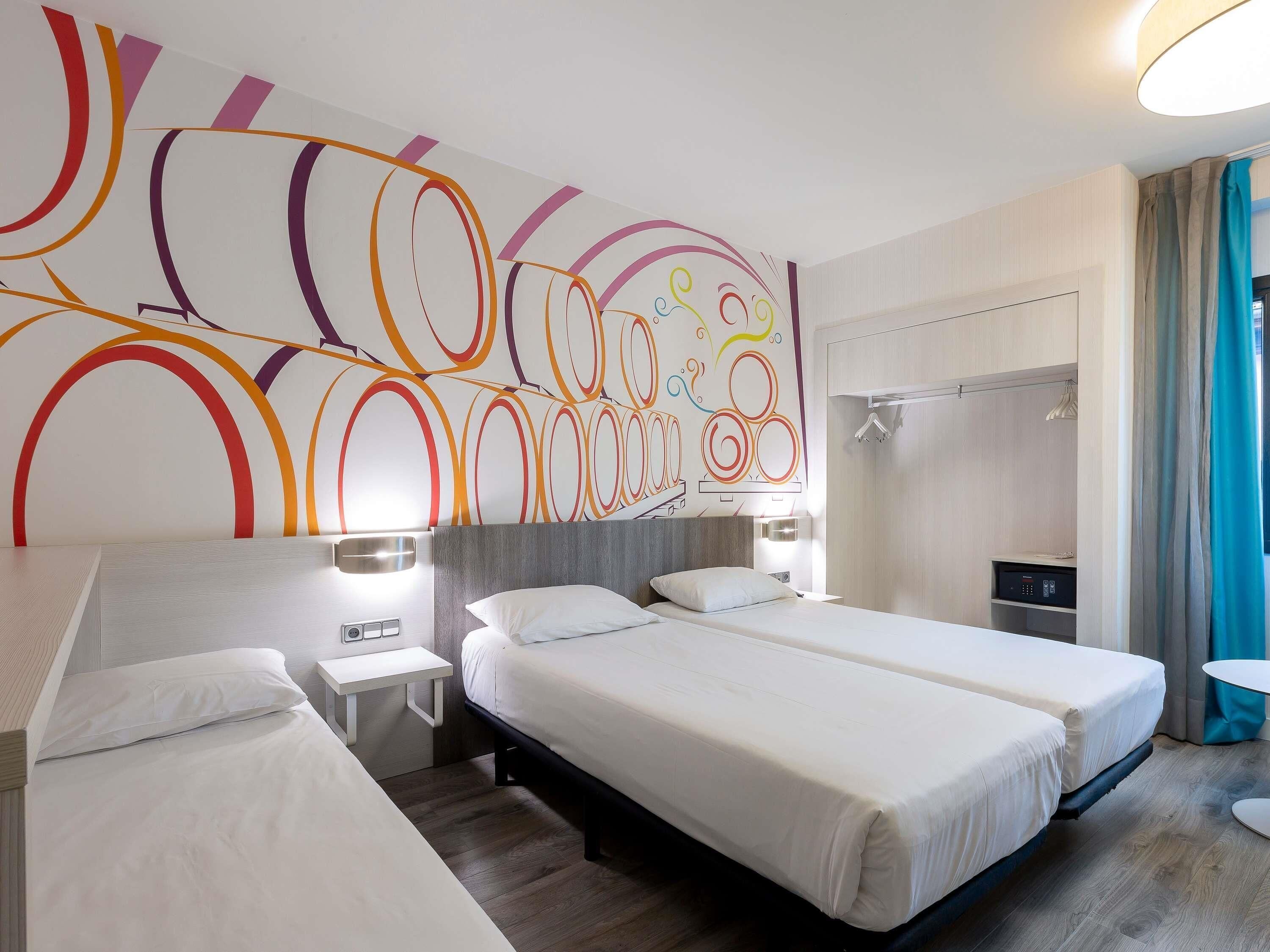 A hotel room with two beds, a modern abstract wall design, a shower area, and a small table.