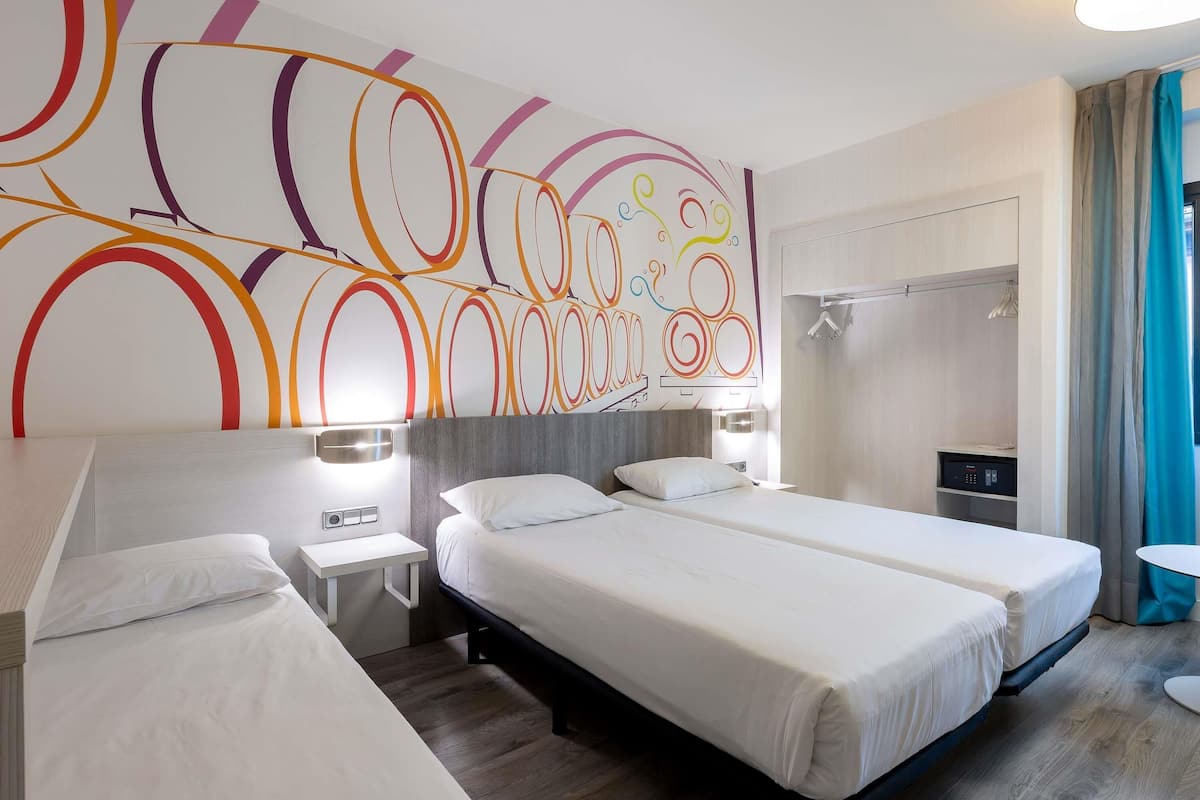 A hotel room with two beds, a modern abstract wall design, a shower area, and a small table.