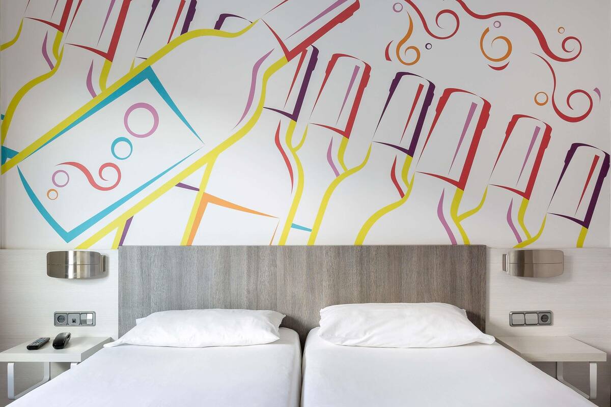 A hotel room with two beds, a wall with colorful abstract art, bedside tables with lamps, and a modern headboard.