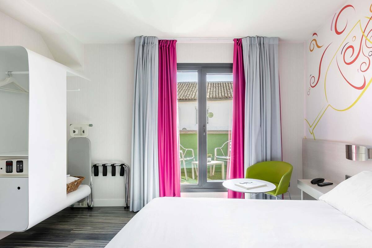 A hotel room with a bed, a small table, a chair, a desk, and a balcony with chairs.