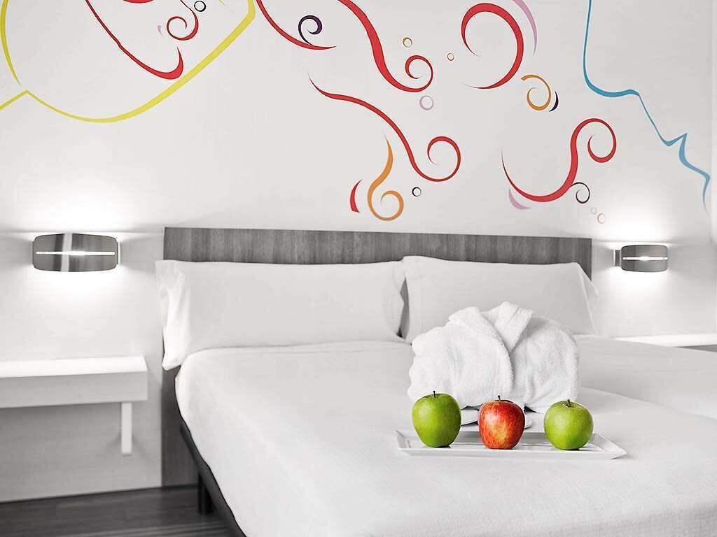 A hotel room with a neatly made bed, a white robe, and two apples on a tray.