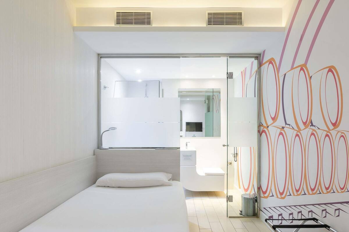 A modern hotel room with a bed, a glass shower enclosure, a sink, and a wall with abstract art.