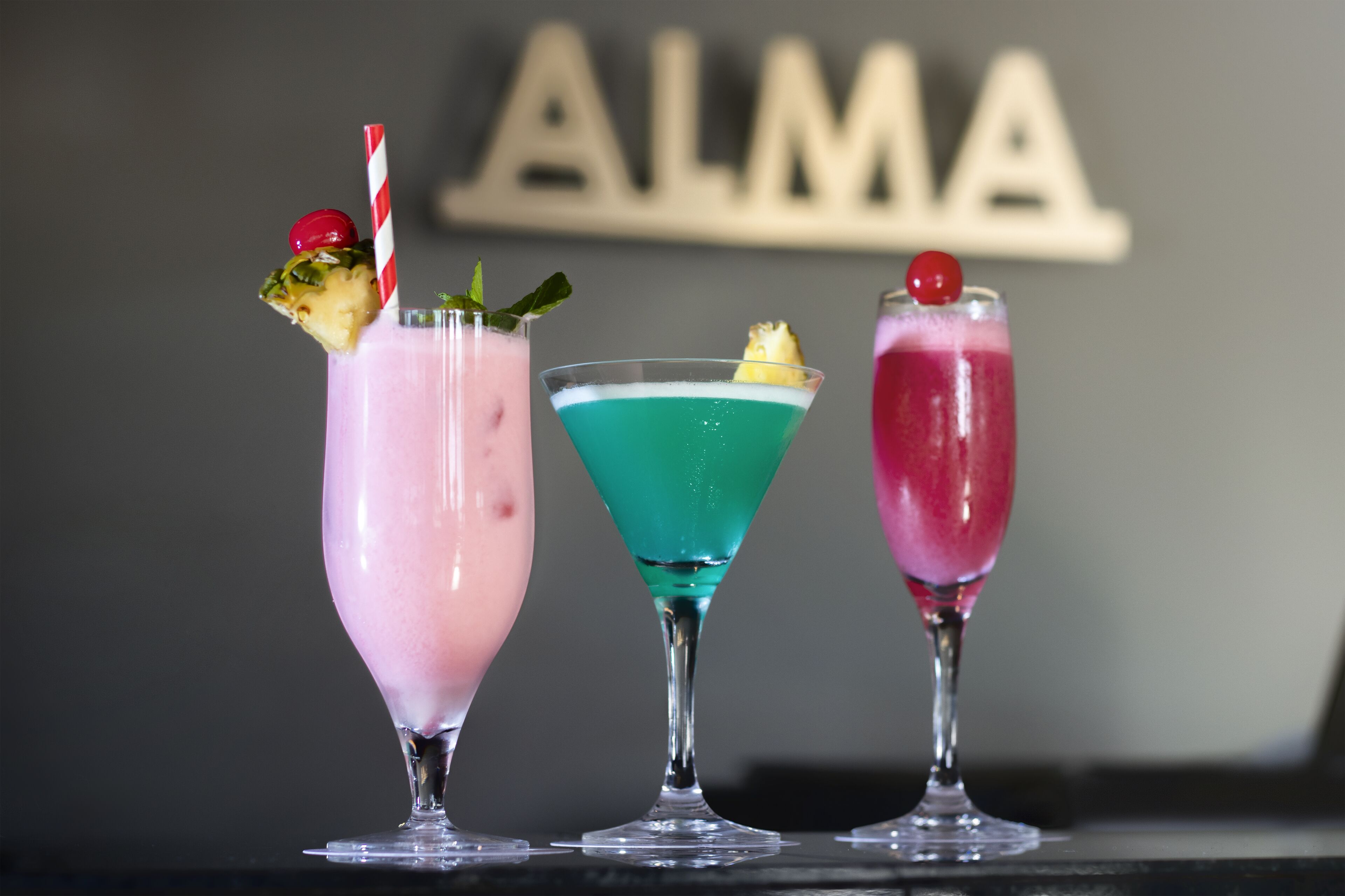 Three cocktails in martini glasses with garnishes, set against a dark background with a sign that reads "ALMA".