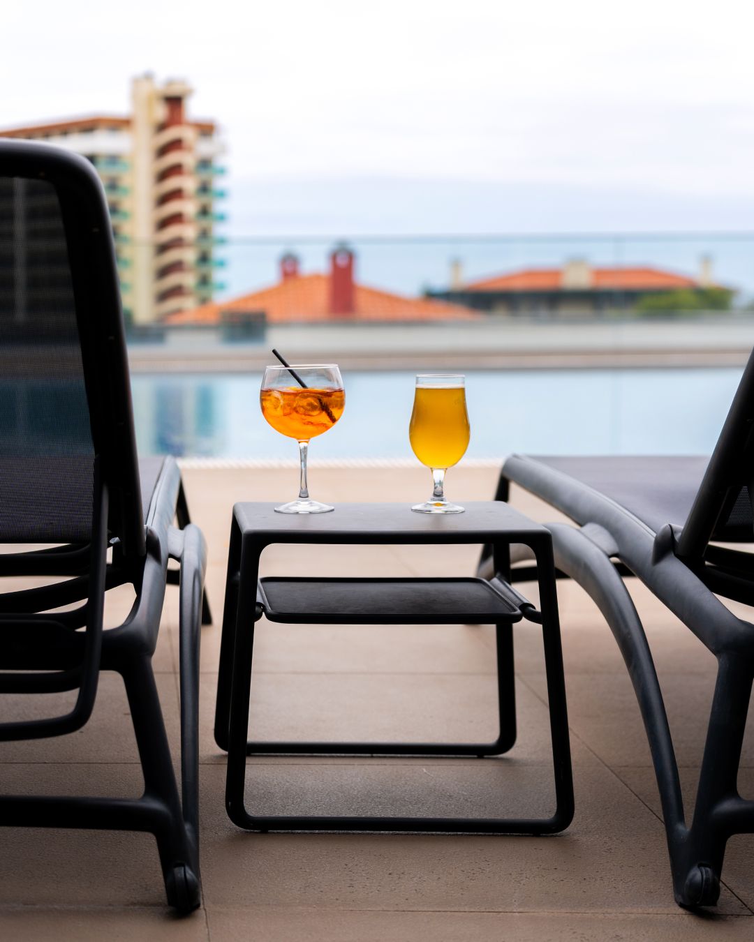 A table with two glasses of beverages on a balcony overlooking a body of water and buildings.