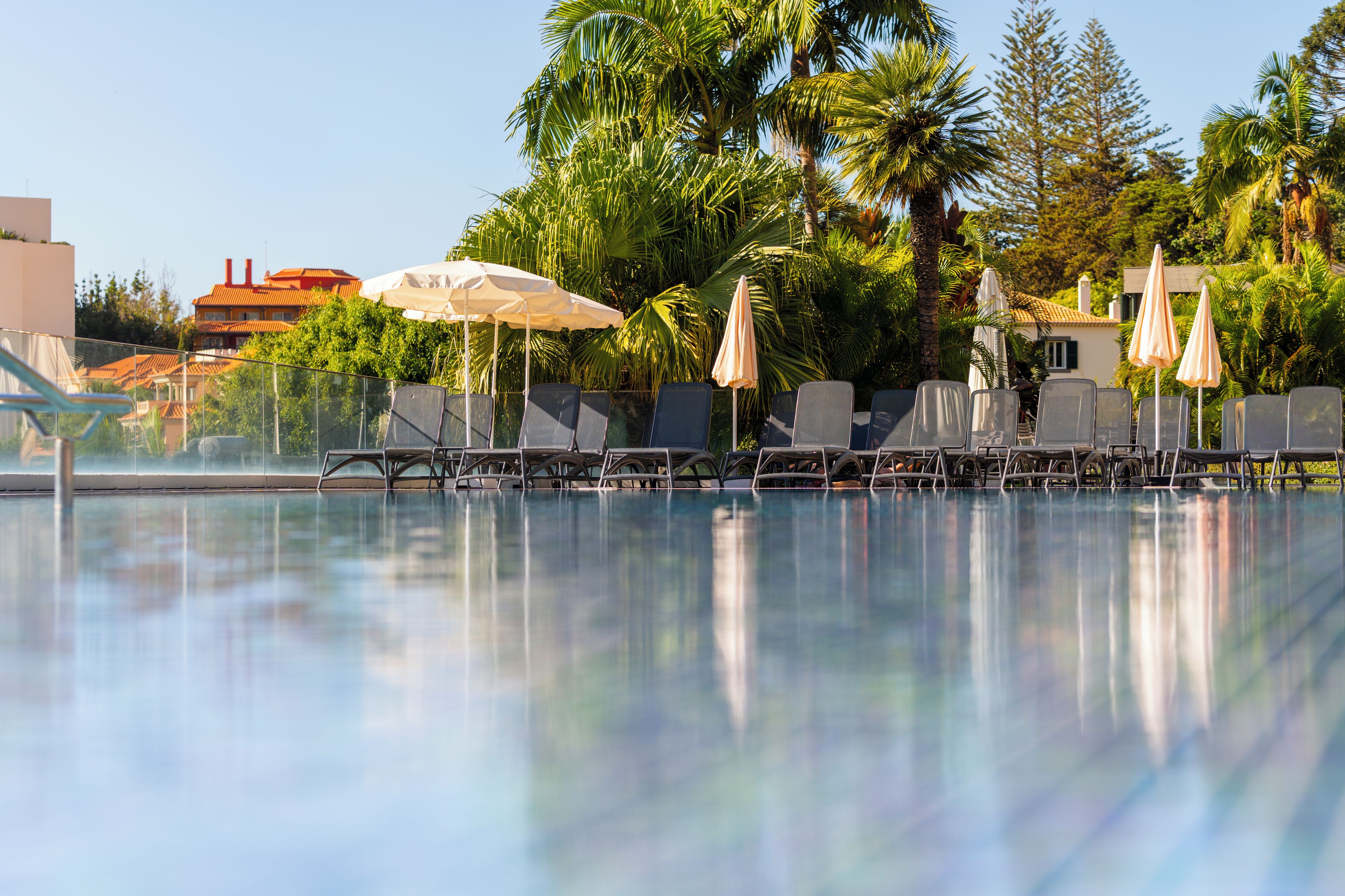 Ein Swimmingpool mit Liegestühlen und Sonnenschirmen, umgeben von Palmen und einem Gebäude mit rotem Dach.
