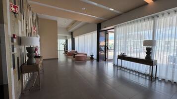 Una hall d'albergo con reception, area relax e ampie finestre con tende leggere.