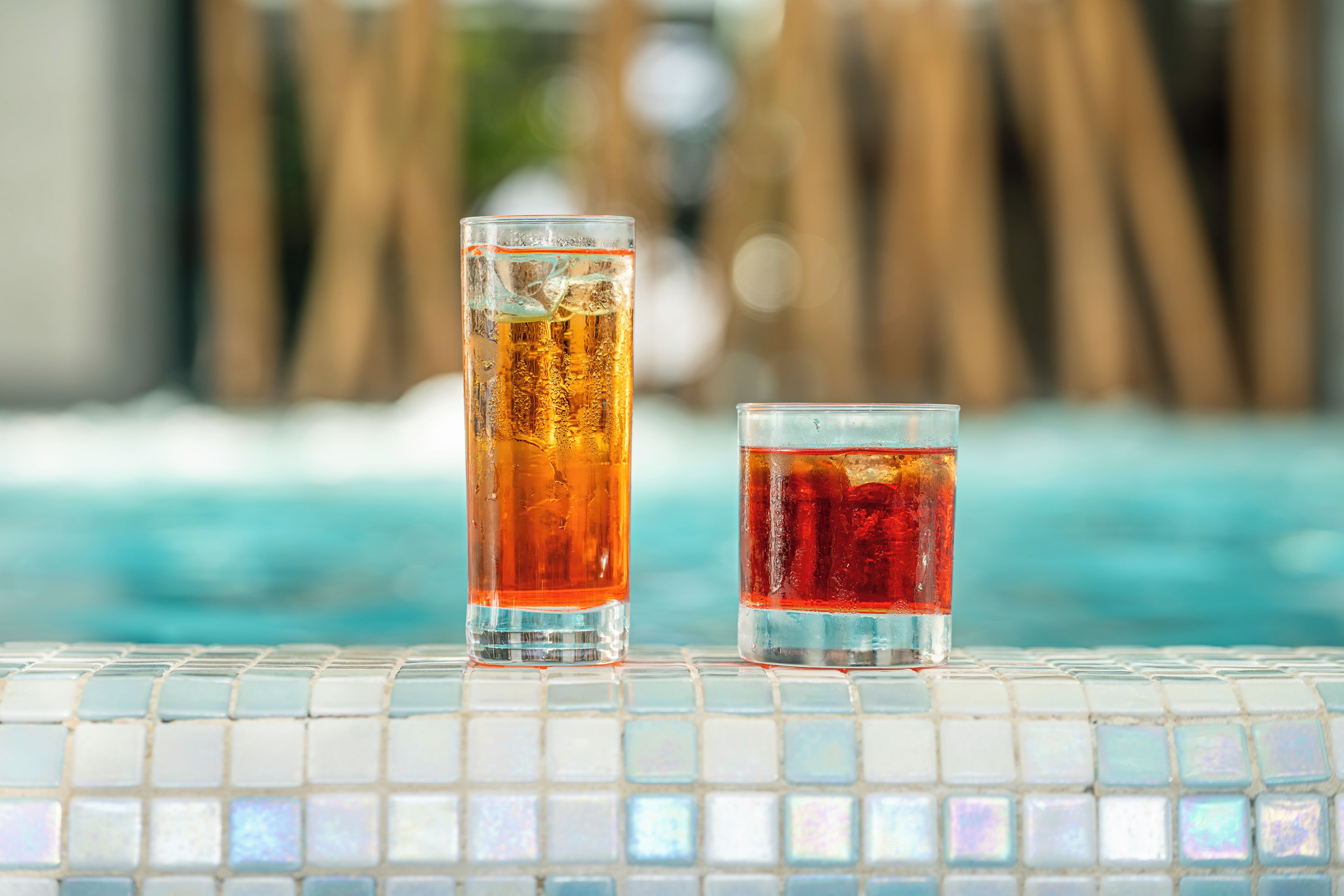 Two glasses of iced beverages on a tiled surface near a pool.