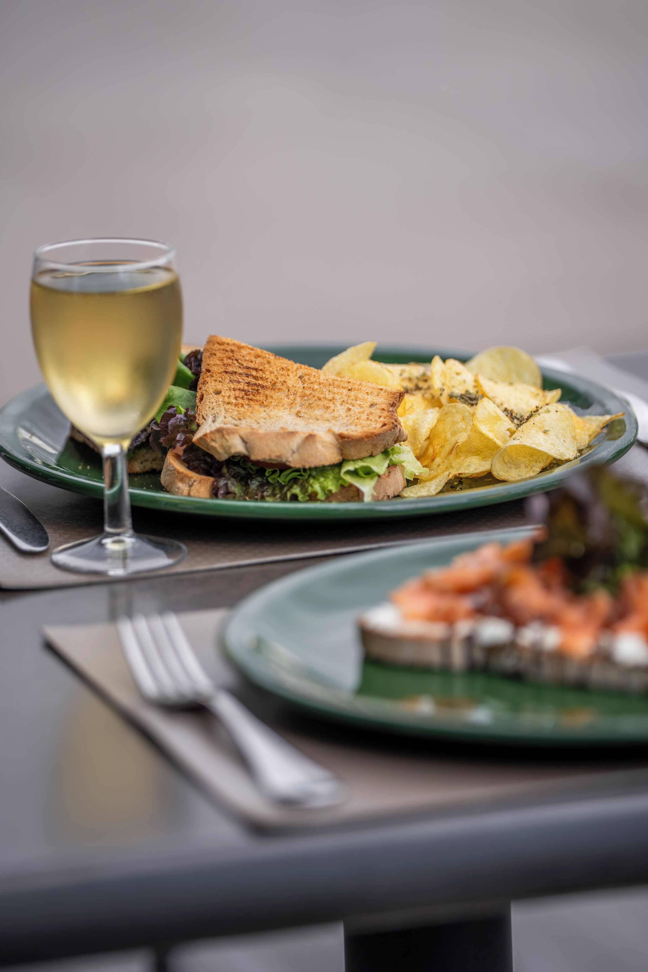 A dining table with two plates of food, one with a sandwich and chips, and a glass of white wine.