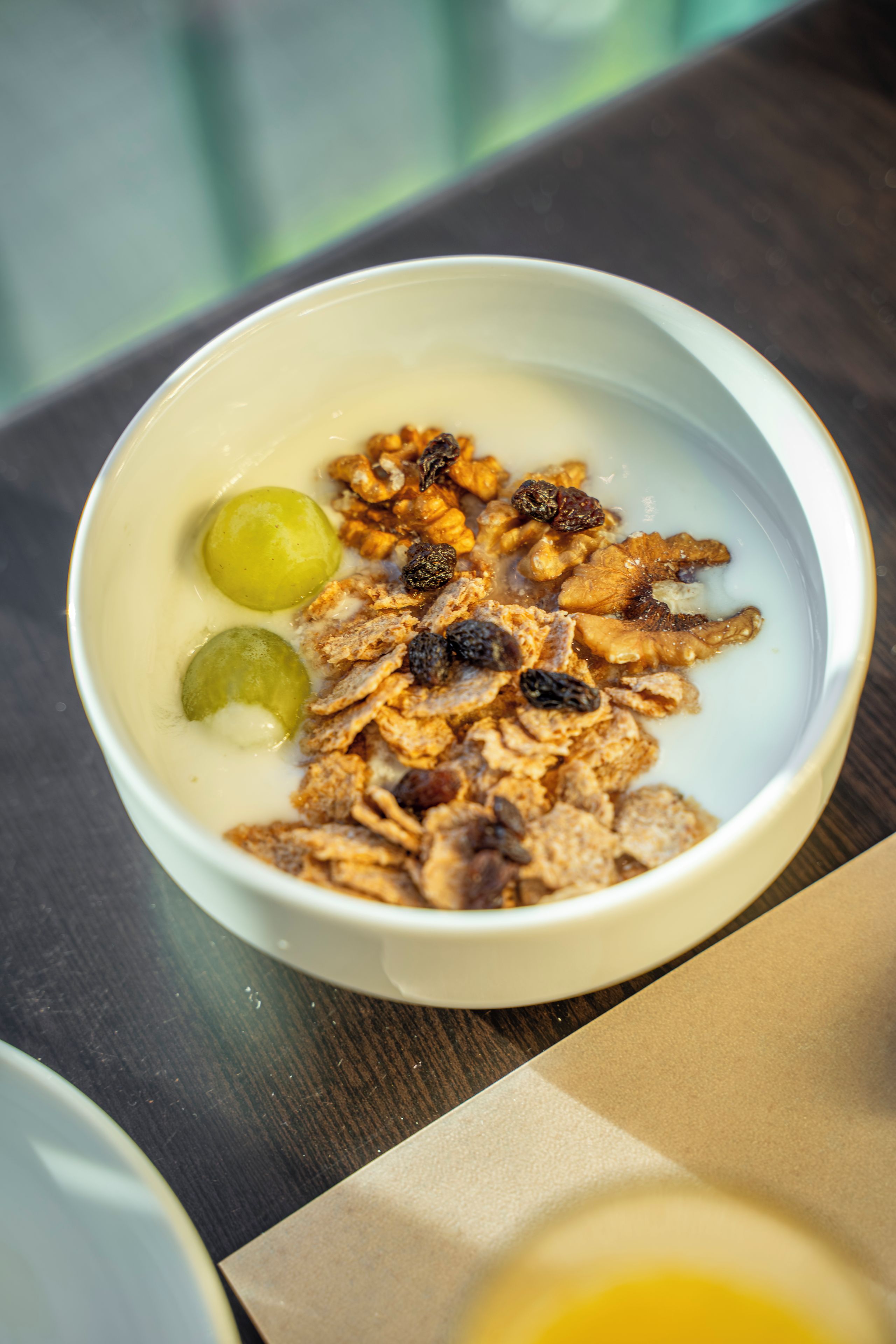 A bowl of cereal with milk, raisins, and grapes on a table.