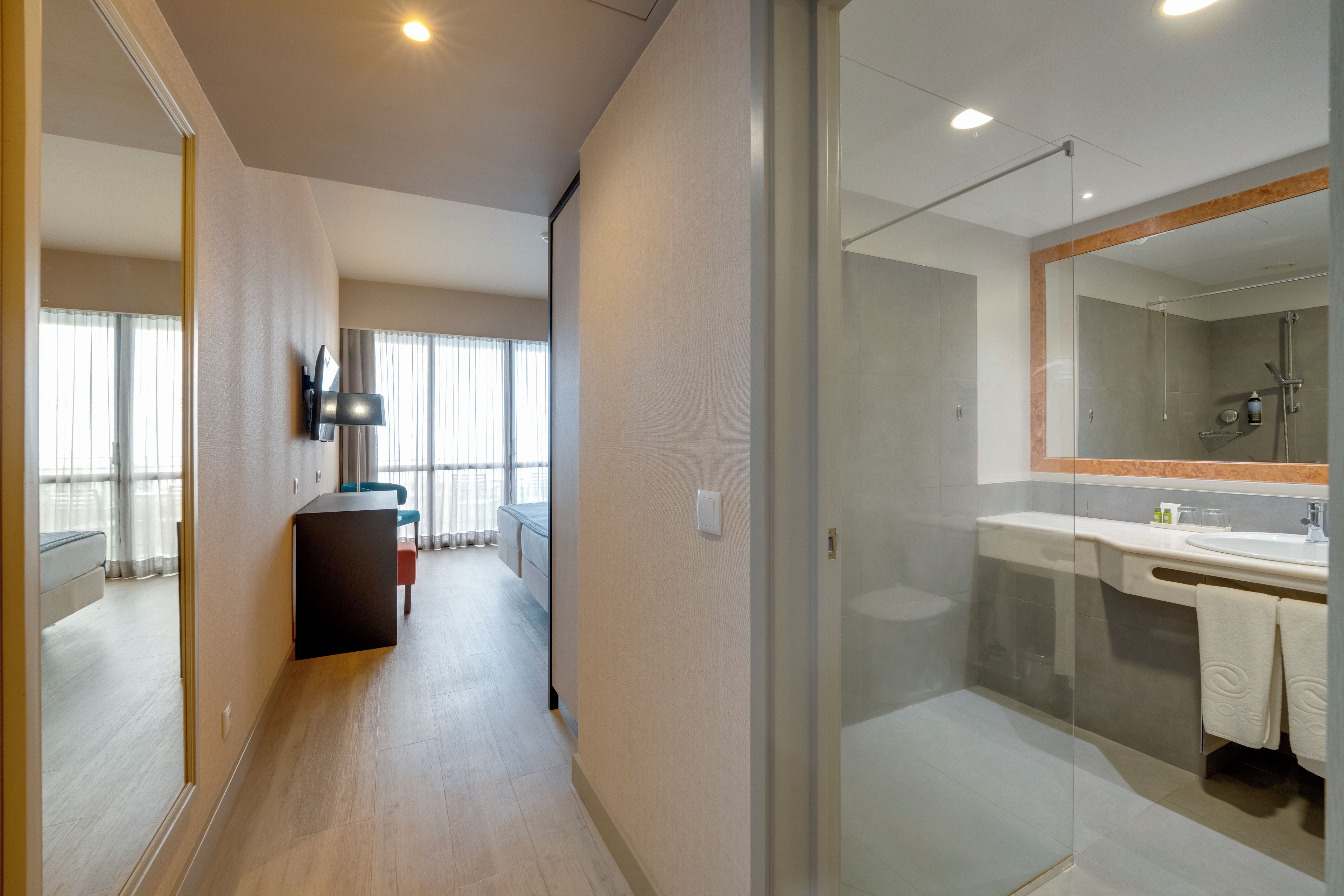 A modern hotel room with a glass shower enclosure, a flat-screen TV mounted on the wall, and a bed with a headboard.