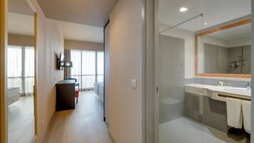 A modern hotel room with a glass shower enclosure, a flat-screen TV mounted on the wall, and a bed with a headboard.