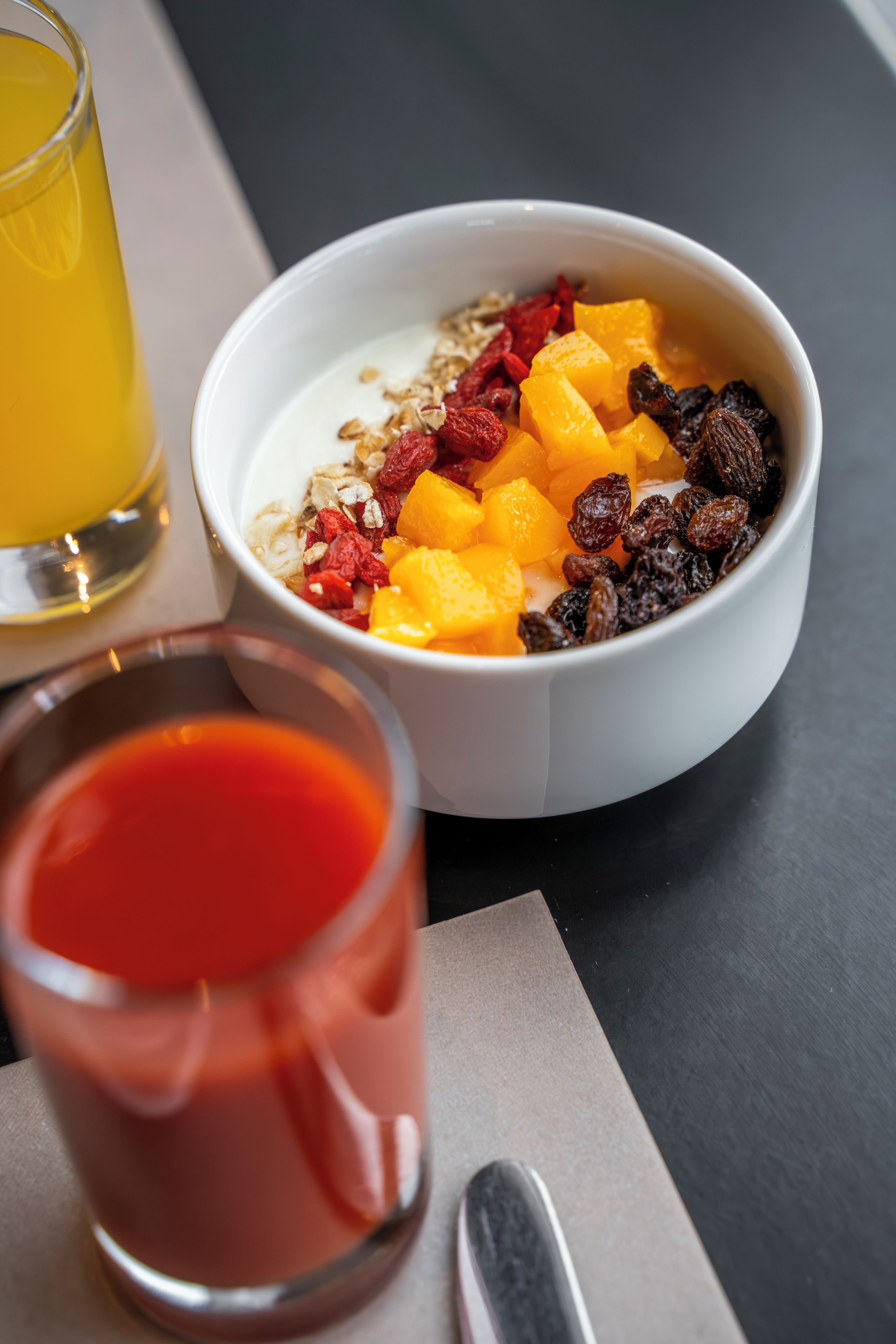 A bowl of cereal with fruits and nuts, a glass of orange juice, and a glass of red beverage on a table.