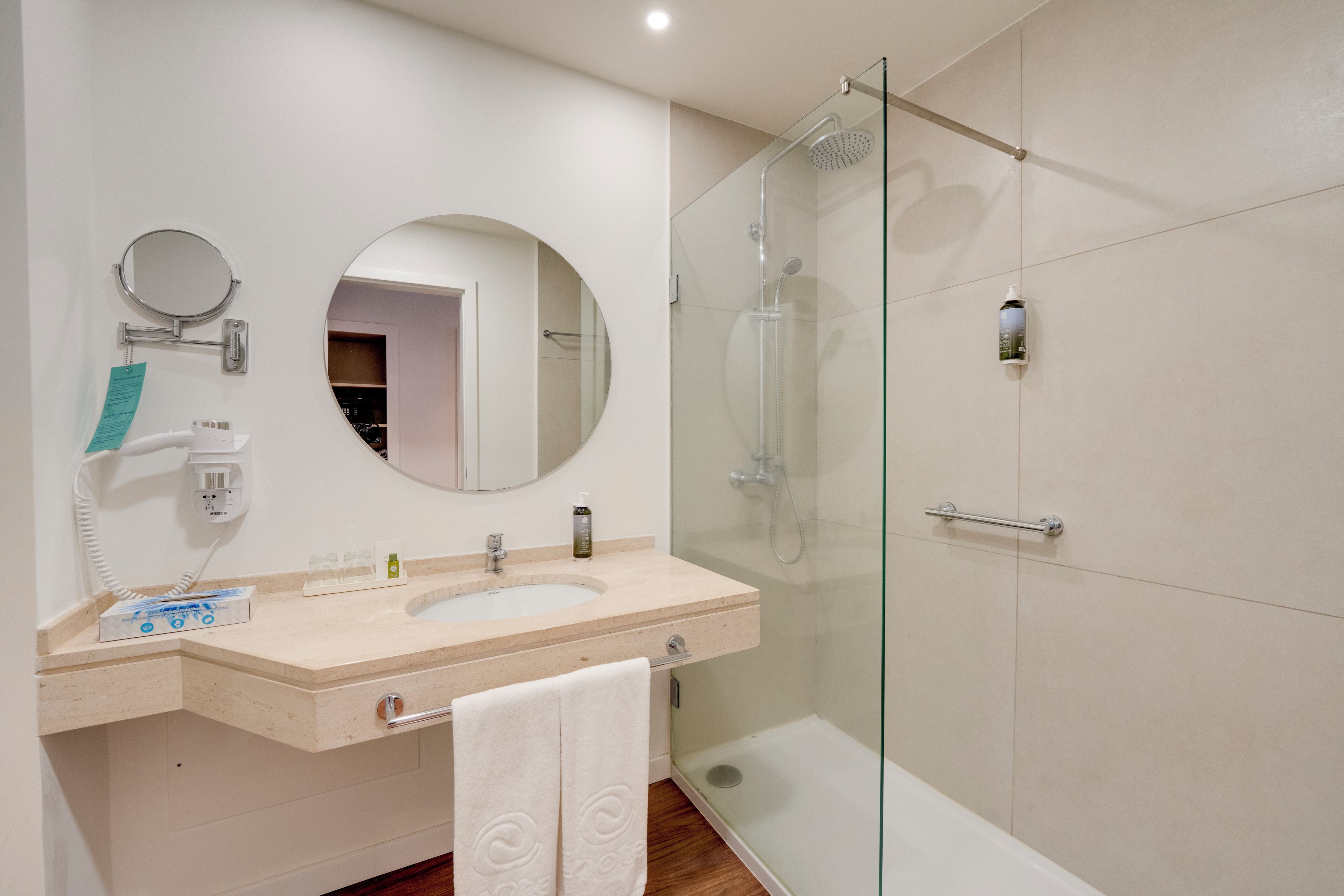 A modern bathroom with a glass-enclosed shower, a round mirror, and a vanity with a towel rack.