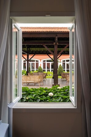 A view through a window of a covered outdoor seating area with a pergola structure, a building with a red-tiled roof in the background, and a garden with green plants and a white flower.