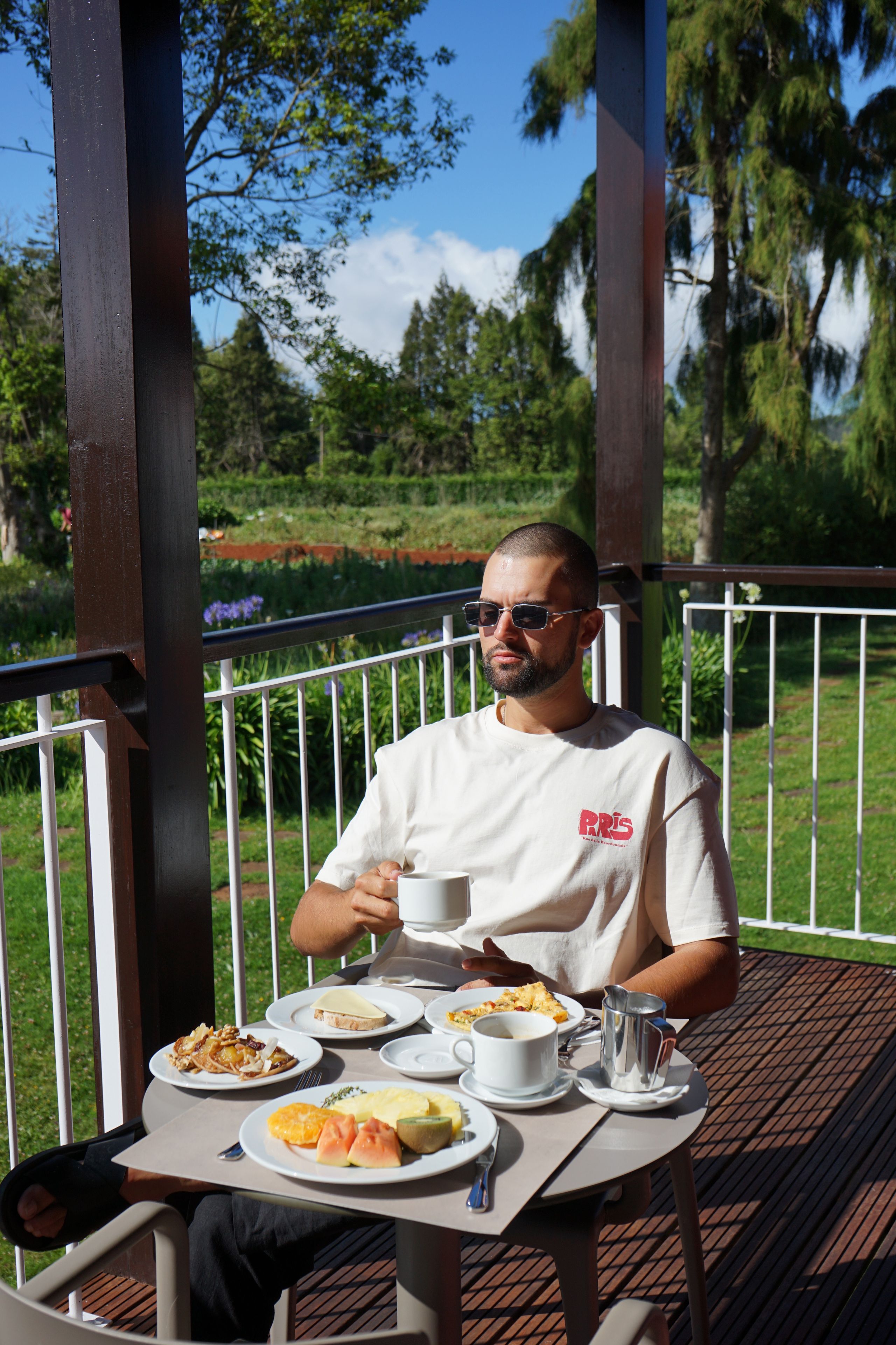 Une personne est assise à une table à l'extérieur, devant des assiettes remplies de nourriture, une tasse et un récipient en métal.