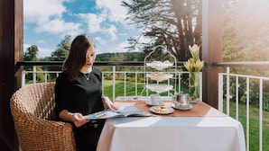 A woman reading a magazine at a table with a three-tiered tea service, a coffee pot, and a vase of flowers on a balcony with a view of a green landscape. - Enotel Santo da Serra (Machico)