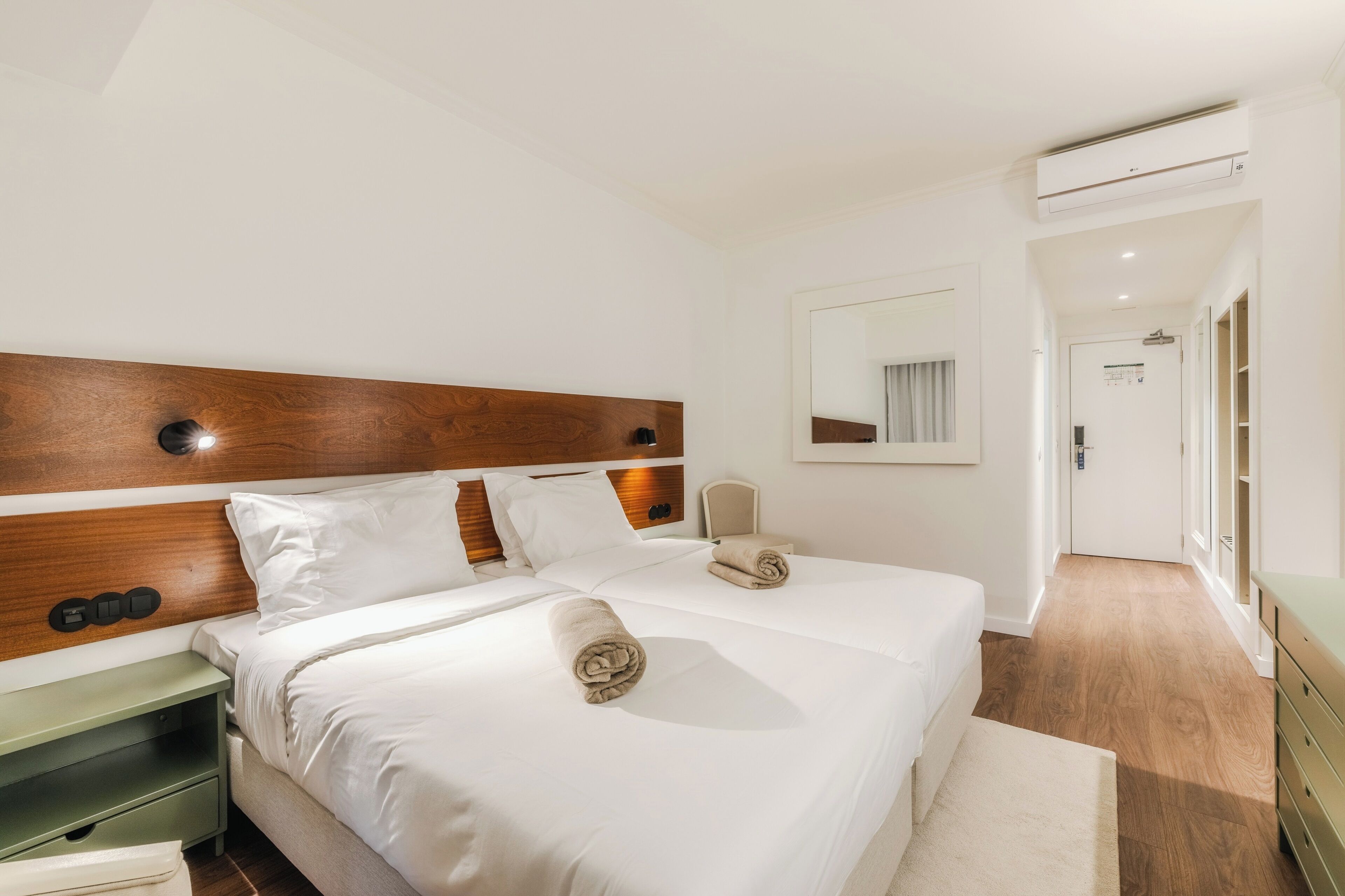 Habitación de hotel con cabecera de madera, cama con ropa blanca, mesita de noche, espejo y equipo de aire acondicionado.