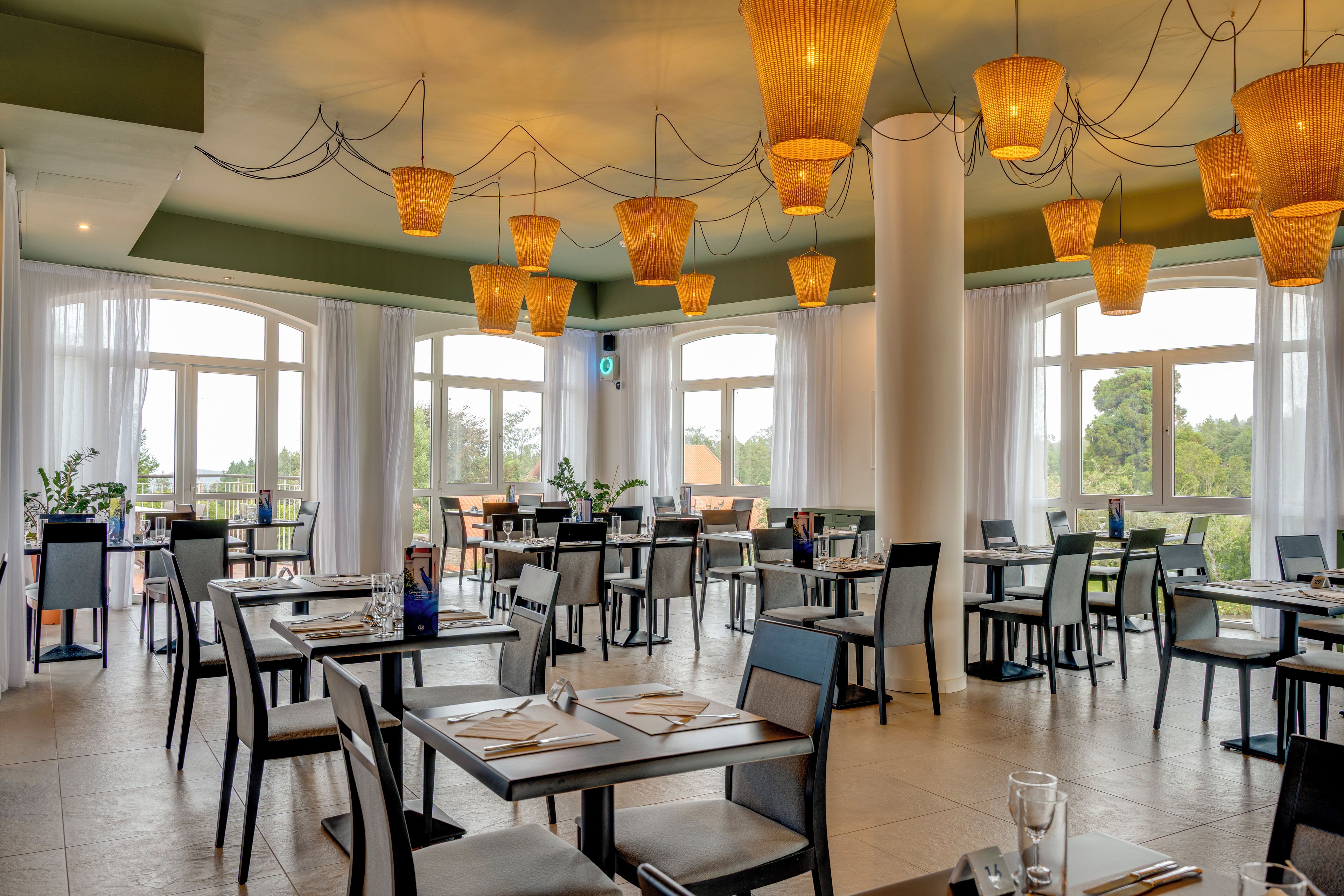 A spacious dining area with multiple tables and chairs, large windows, and hanging wicker chandeliers.