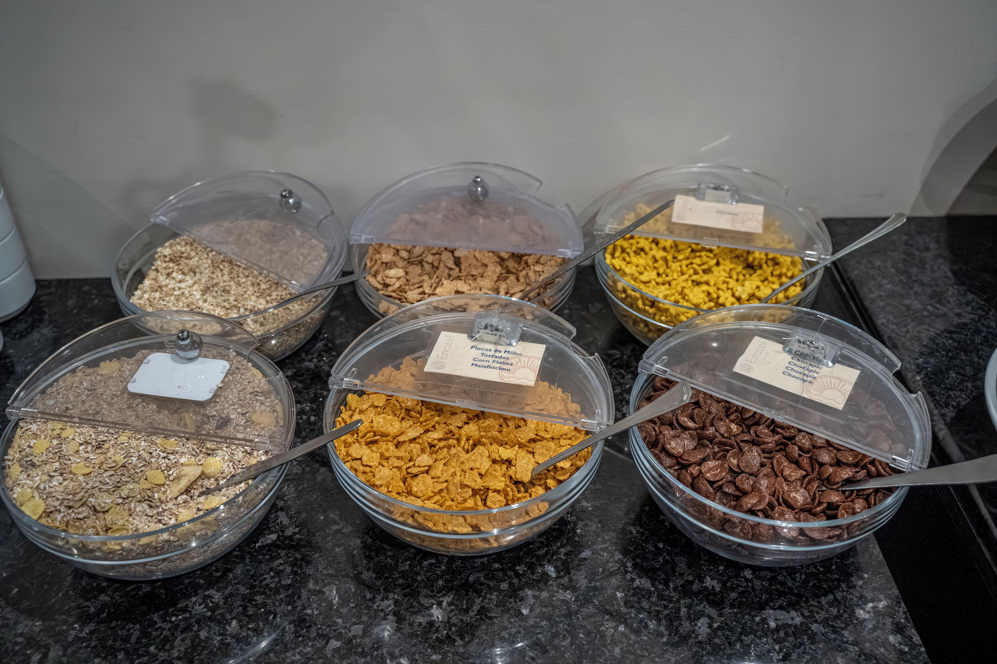 Six clear plastic containers with labels, each containing different types of cereal on a countertop.