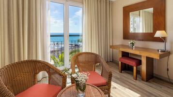 Quarto de hotel com varanda, cadeiras de vime, uma mesa pequena, escrivaninha de madeira e um espelho.