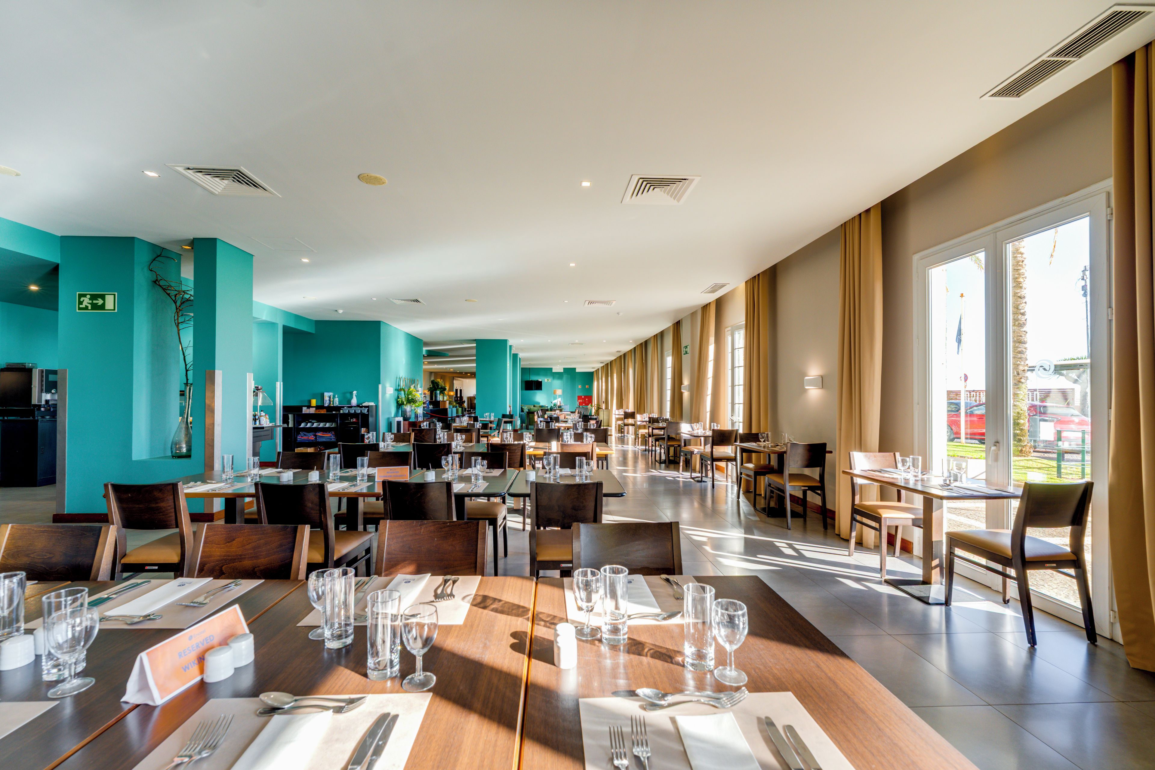 A spacious dining area with multiple tables set for guests, a kitchen area in the background, and large windows allowing natural light.