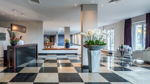 Un vestíbulo de hotel con piso a cuadros, una recepción, una maceta grande con flores blancas y un área de estar con un sofá y sillones.