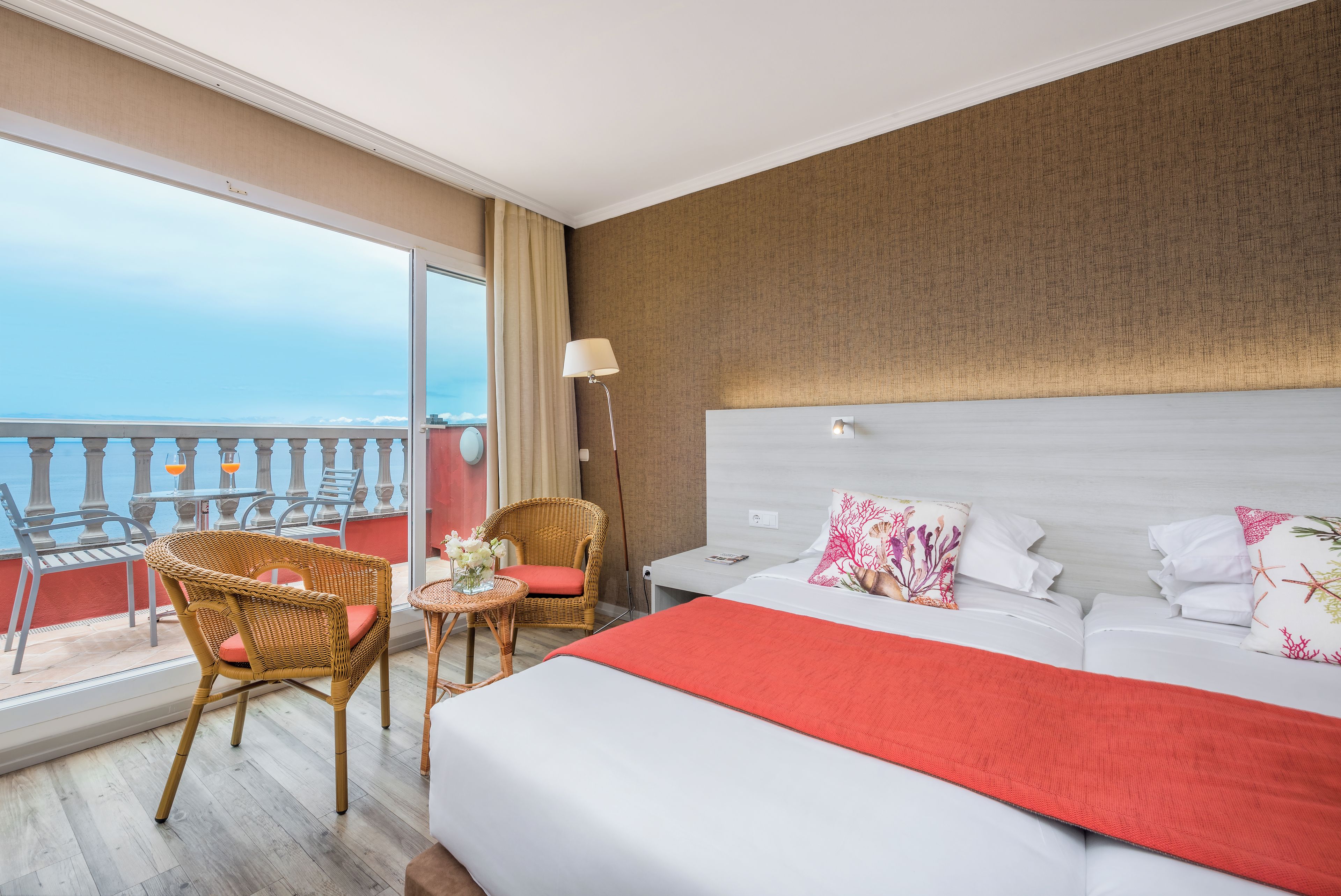 A hotel room with a balcony, a bed with white bedding and a red throw, a wicker chair with a cushion, and a small table with a vase of flowers.