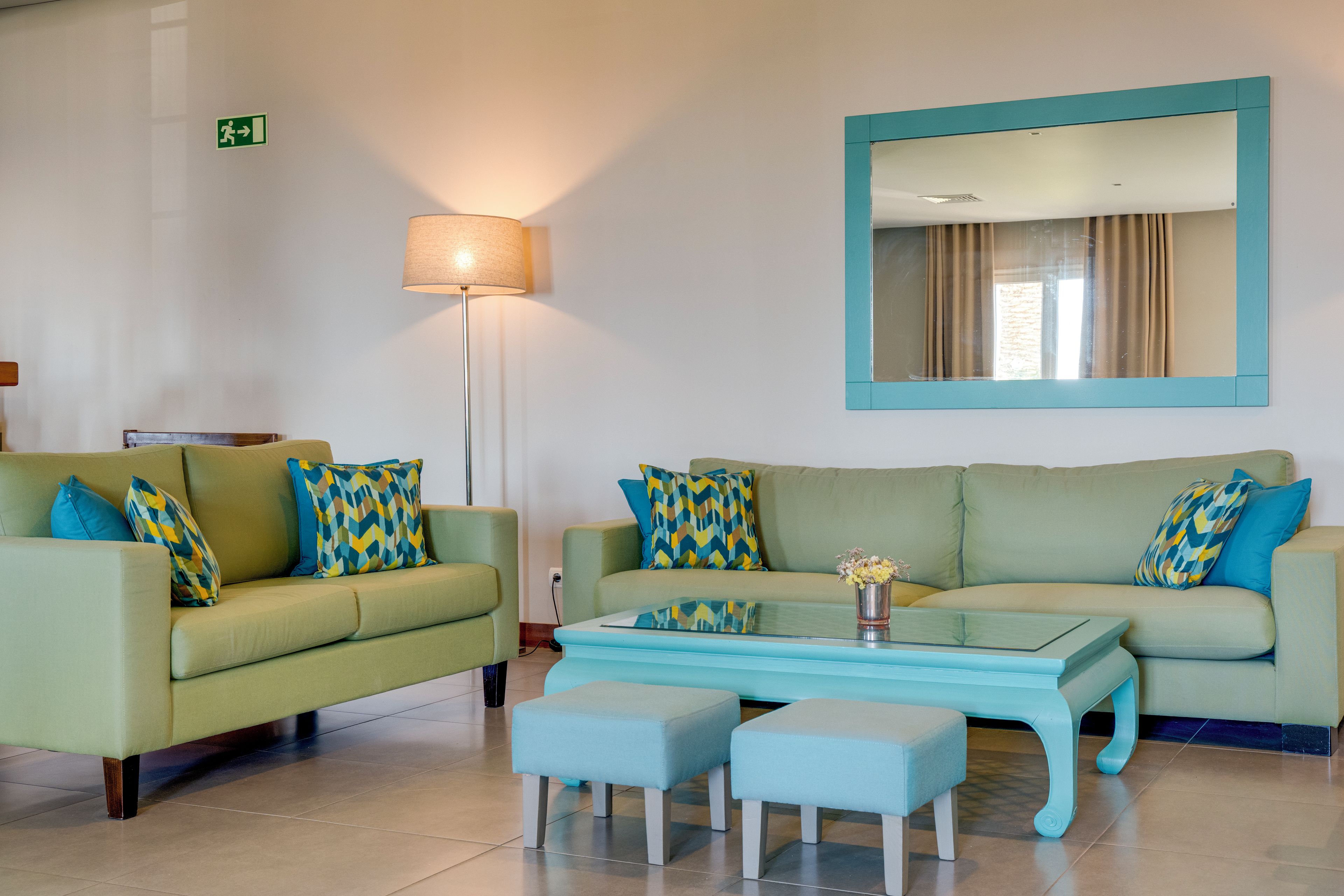 A hotel room with two green sofas, matching ottomans, a glass coffee table, a floor lamp, and a large mirror on the wall.